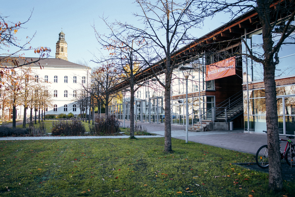 Bild zeigt großes Gebäude, davor eine Grünfläche mit Bäumen.