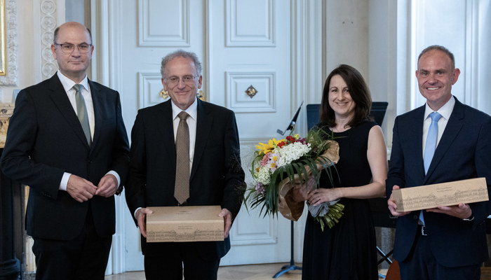 4 Personen, eine Frau und drei Männer in einem festlichen Saal. Die Frau hält einen Blumenstrauß, zwei der Männer halten ein Geschenkpaket.